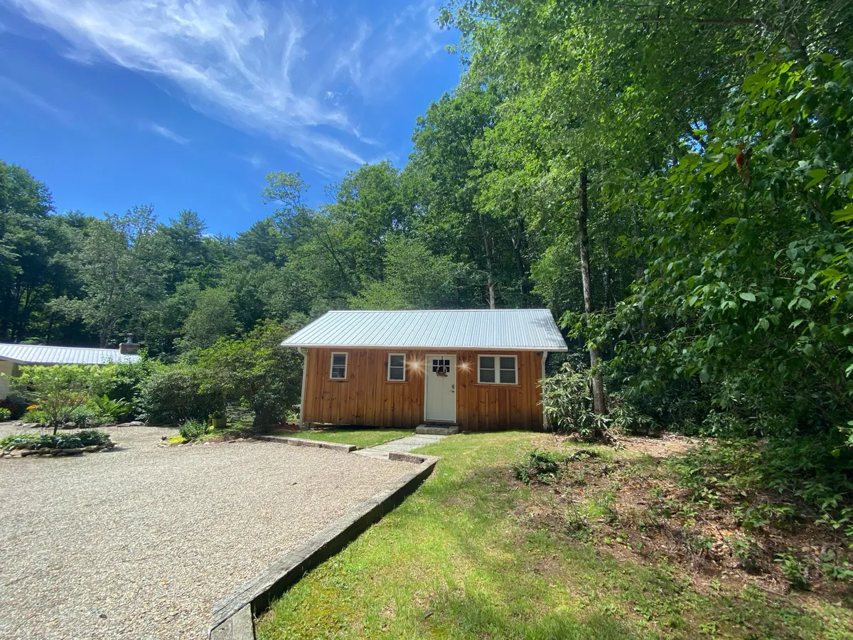 The image shows a charming, compact wooden cabin with a sleek metal roof, nestled among lush, mature trees. A gravel driveway leads up to the cabin, and the serene forest setting creates a sense of peaceful seclusion. The porch hints at a perfect spot to relax and watch sunsets, while the overall scene feels cozy and inviting.