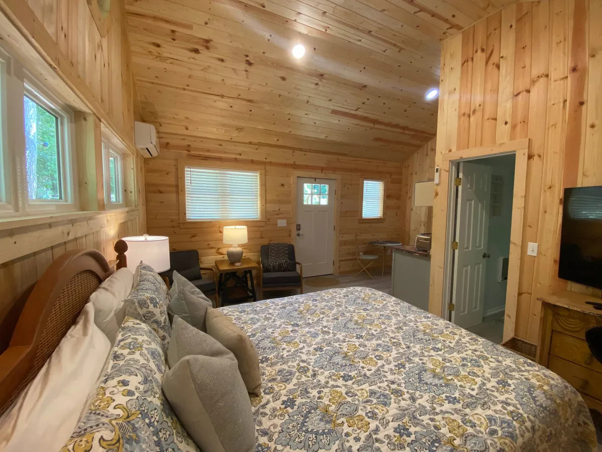 A cozy interior with a bed dressed in patterned bedding sits alongside an armchair and lamp. The wooden walls and ceiling create a warm, enveloping feeling, while the arrangement encourages relaxation, reading, or evening conversations by soft lamp light.