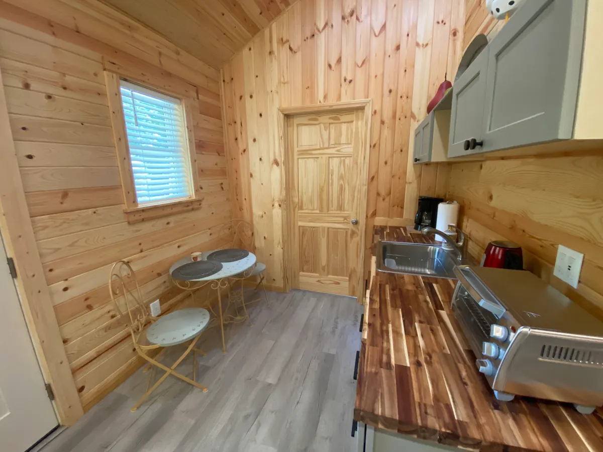 A warm interior scene with wooden walls, a table and two chairs set beside kitchen appliances and cabinetry. Morning light streams through a window, illuminating the inviting workspace. The setup is intimate and functional, ideal for working, reading, or enjoying a quiet coffee.