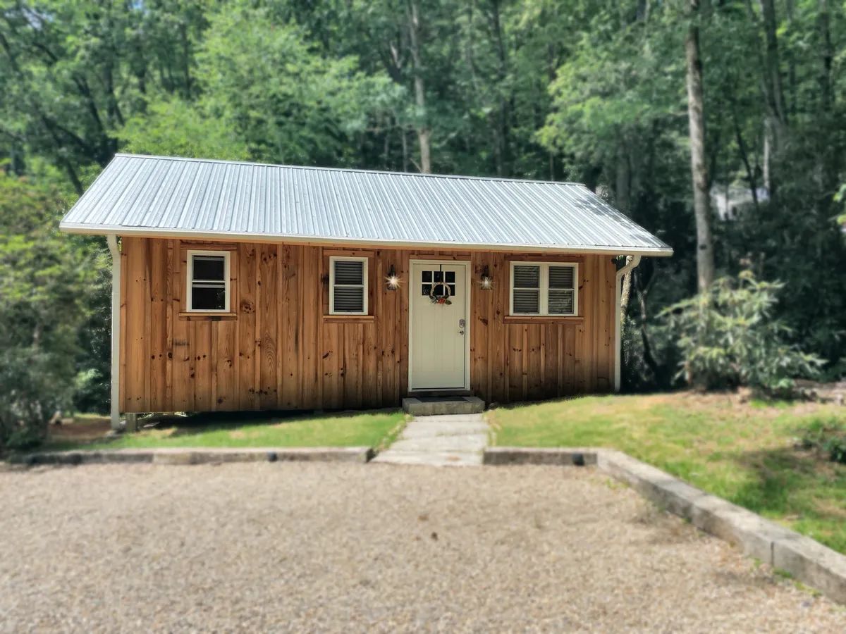 A cozy, welcoming exterior view of the suite's wooden facade, featuring a private front door and a landscaped entrance. The setting evokes a sense of privacy and tranquility while hinting at convenient access, with the porch and immediate outdoor space inviting morning relaxation.