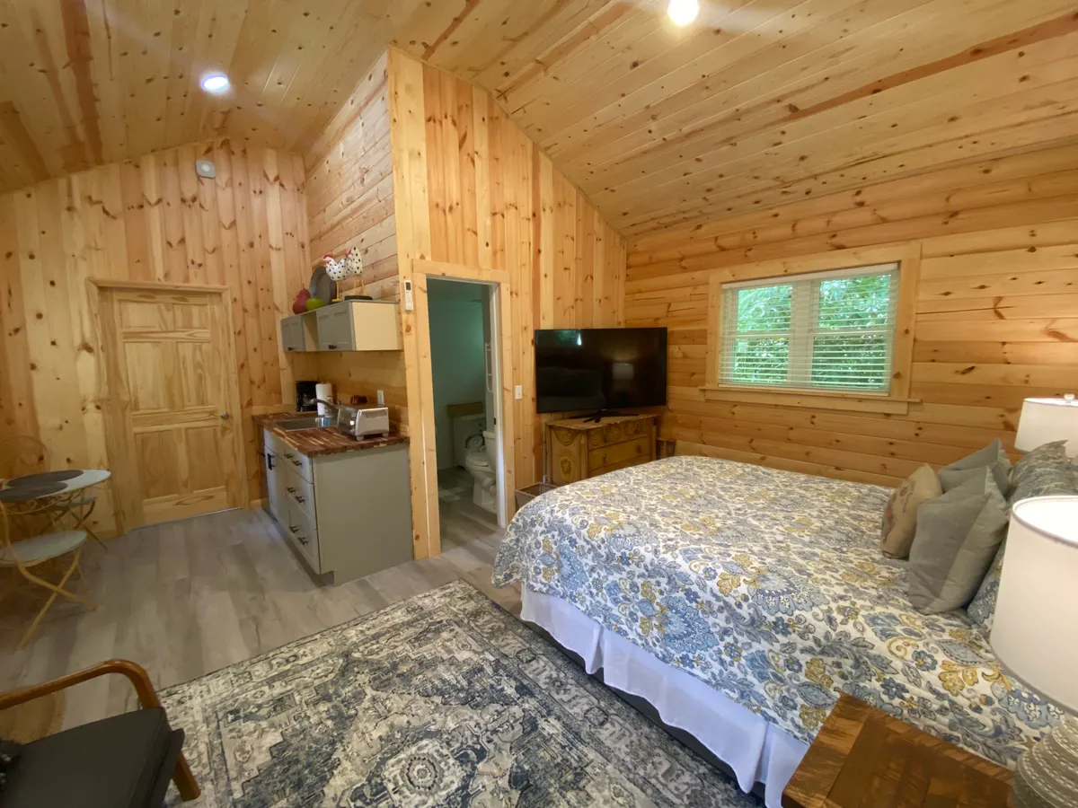 This interior shot centers on a bed with floral bedding against warm wooden wall paneling. A small kitchen area and a television set are visible, with a decorative rug anchoring the space. A table and chair are partially visible off to the side, suggesting a multi-use area for both relaxation and work. Soft, natural light fills the room, enhancing the cozy and adaptable atmosphere.