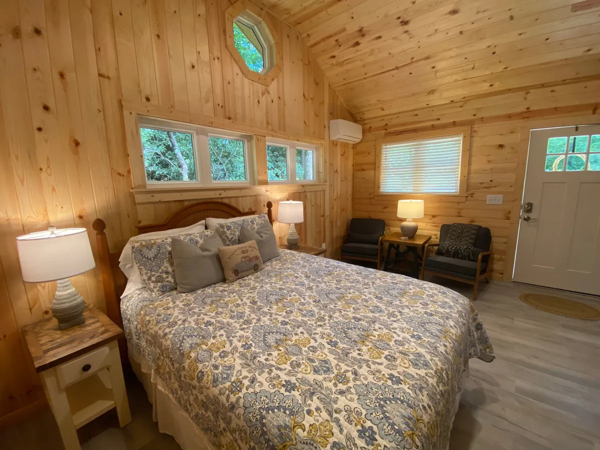 This image displays a warmly lit bedroom featuring a plush bed with floral bedding, wooden walls, windows allowing natural light to gently pour in, side tables with lamps, and inviting armchairs in the corner. The tranquil décor and soft lighting foster a welcoming, restful atmosphere.