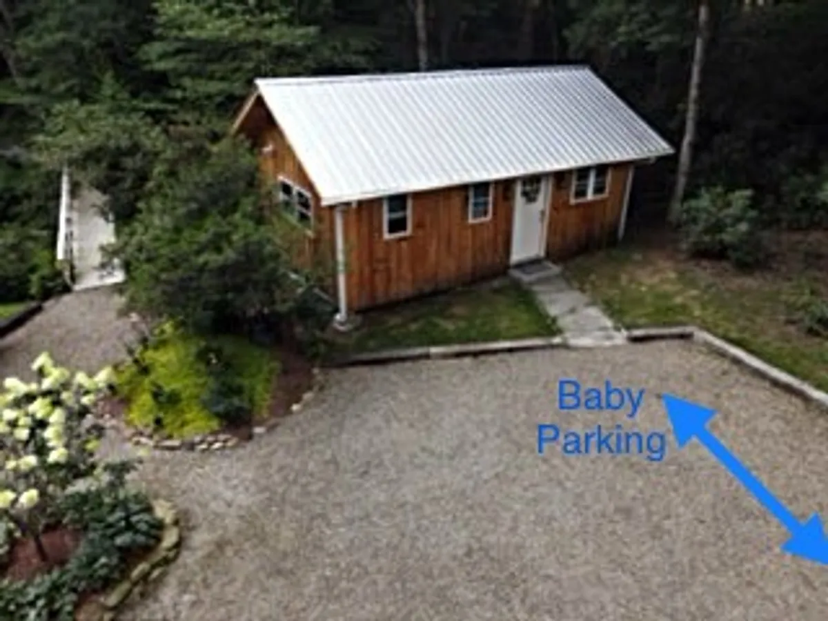 A wide exterior shot of a wooden cabin with a clearly visible gravel parking area directly adjacent to the property, surrounded by a landscaped garden. The image shows the ease and proximity of parking, as well as the connection between the parking area and the private entrance.