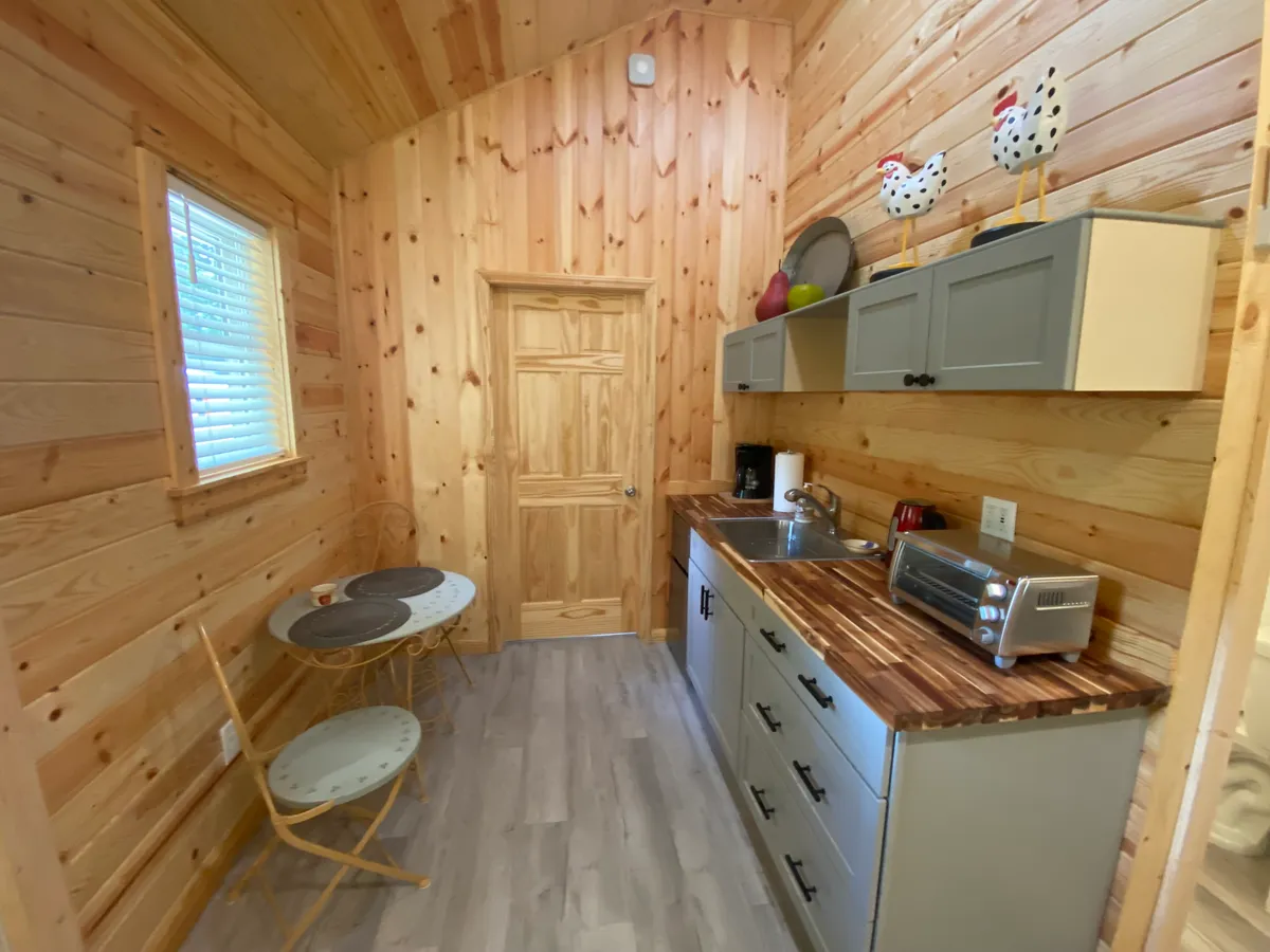 A warm, inviting kitchenette area with wooden cabinetry, a kitchen sink, decorative chicken accents, and a small dining table set for a meal. The space feels homey and functional, perfectly suited for light meal preparation and cozy breakfasts.